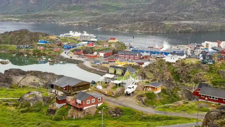 Aerial view of Sisimiut Greenlandic town with colorful buildings, green hills & Nat Geo small ship docked along the shore.
