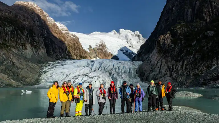 Chilean Patagonia cruise travelers in bright coats stand on stony shore in front of an icy blue glacier & snowy peaks behind.