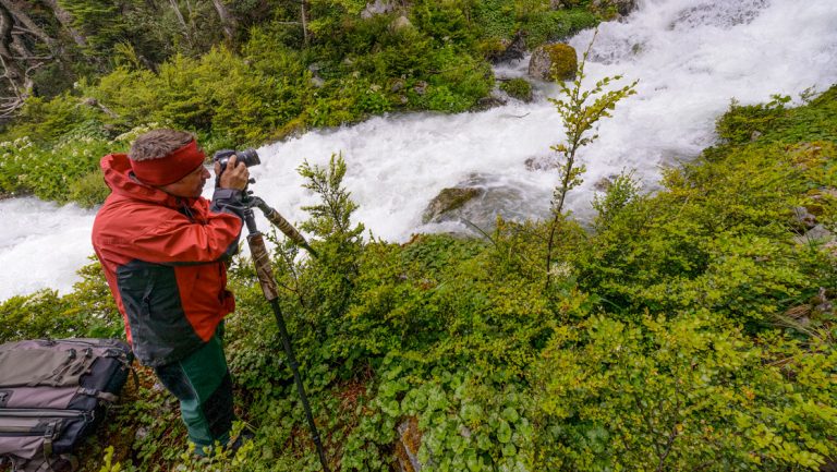 Man in red jacket & headband uses tripod to photograph rushing river below while standing in lush green undergrowth in Patagonia.