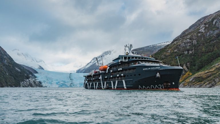 Magellan Explorer dark blue small ship sits in calm water by blue glacier on cloudy day in Patagonia.