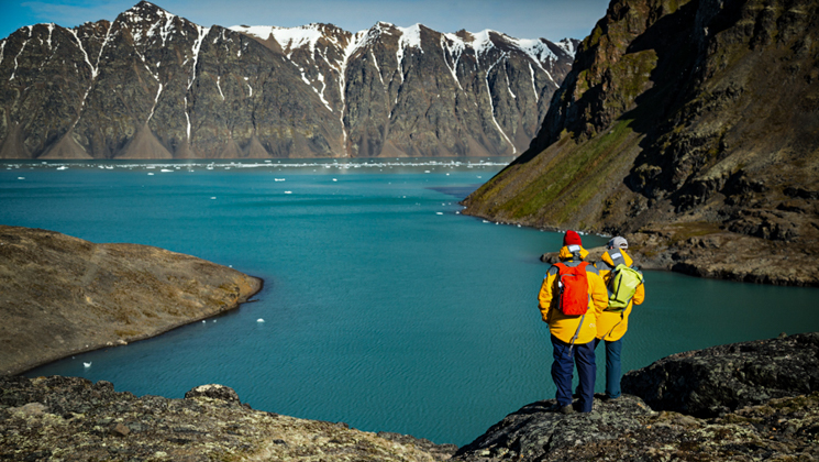 2 Ultimate Arctic Voyage travelers in yellow coats stand on cliff overlooking turquoise fjord with snowcapped mountains behind.