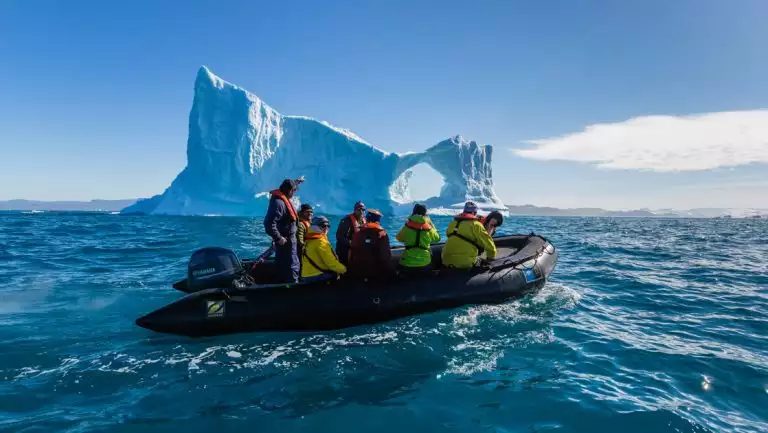 Black Zodiac boat with Greenland travelers in colorful jackets cruises past a large iceberg with a circle cut out.