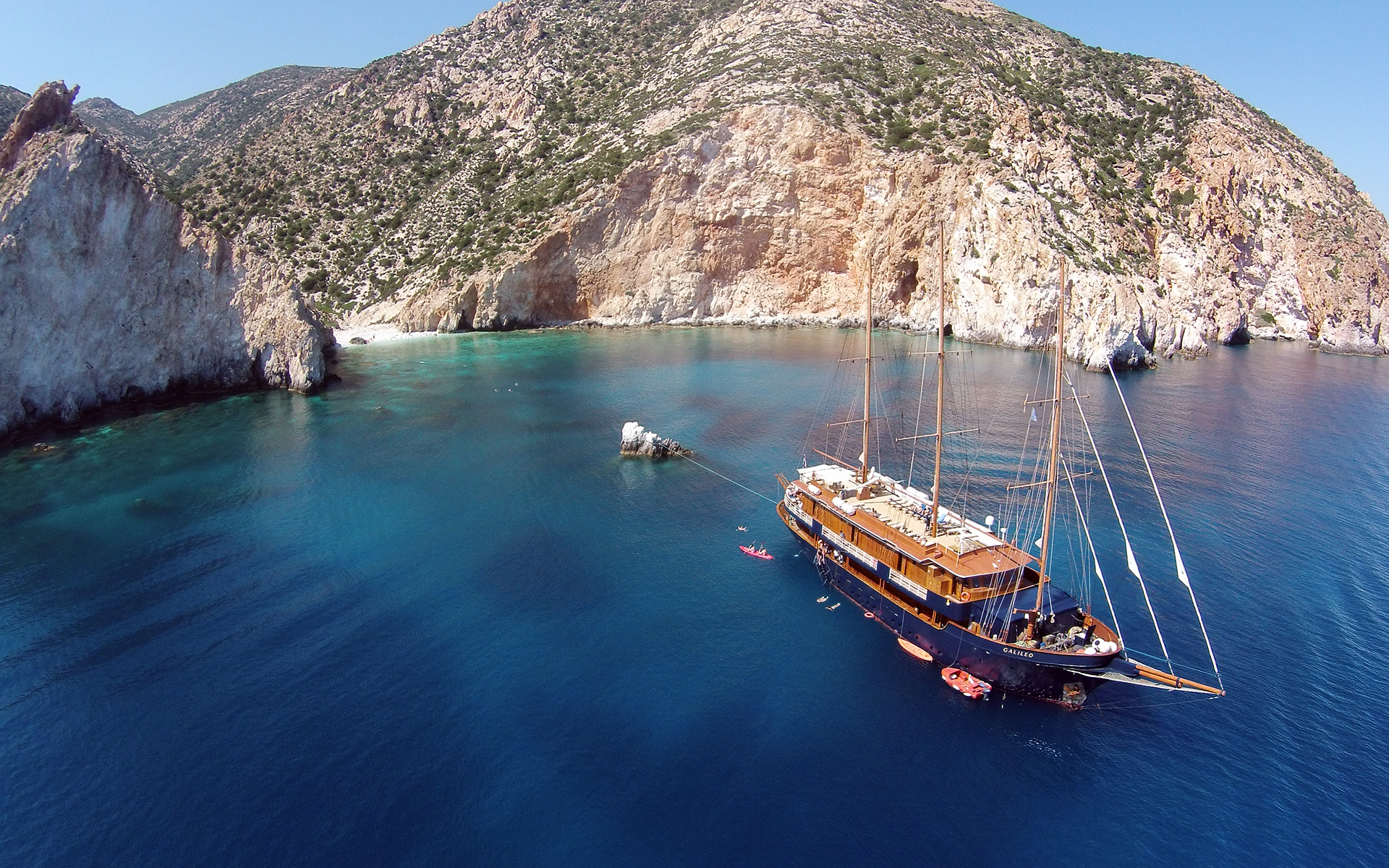 A small sailing ship is seen at anchor in the Mediterranean Sea in a remote cove with tall cliffs and a white beach
