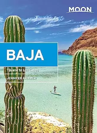 Book cover of the Moon Guide to Baja California by Jennifer Kramer with two large green cactus framing a person on a paddle board in a green water bay with blue sea and brown mountains with blue sky and white clouds. 