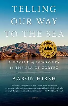 Book cover of Telling Our Way to the Sea by Aaron Hirsh showing brown desert mountains of Baja rising out of a calm blue sea and blue sky above. 