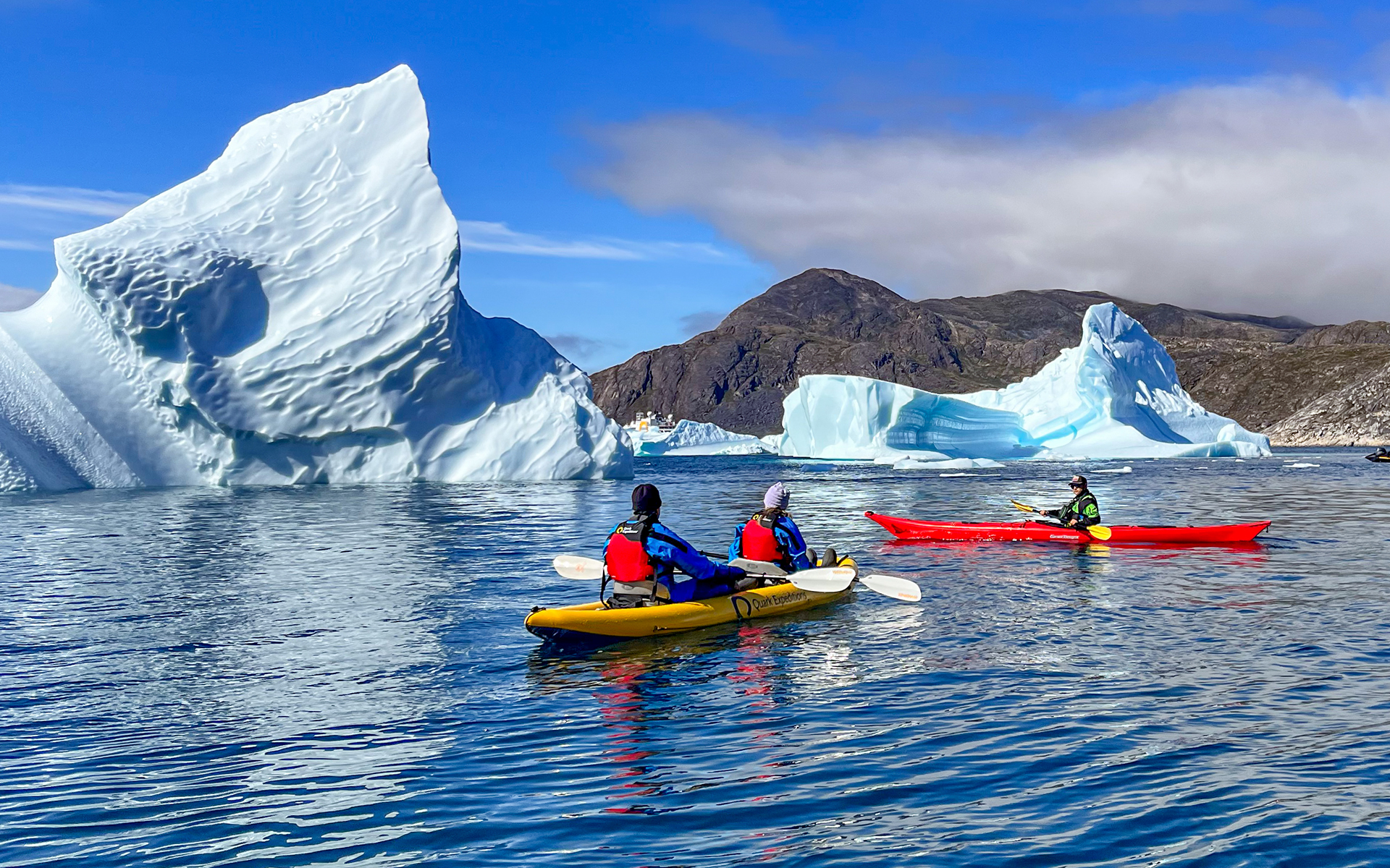On a sunny day a group of kayakers paddle brightly colored red and yellow kayaks among jagged giant floating icebergs