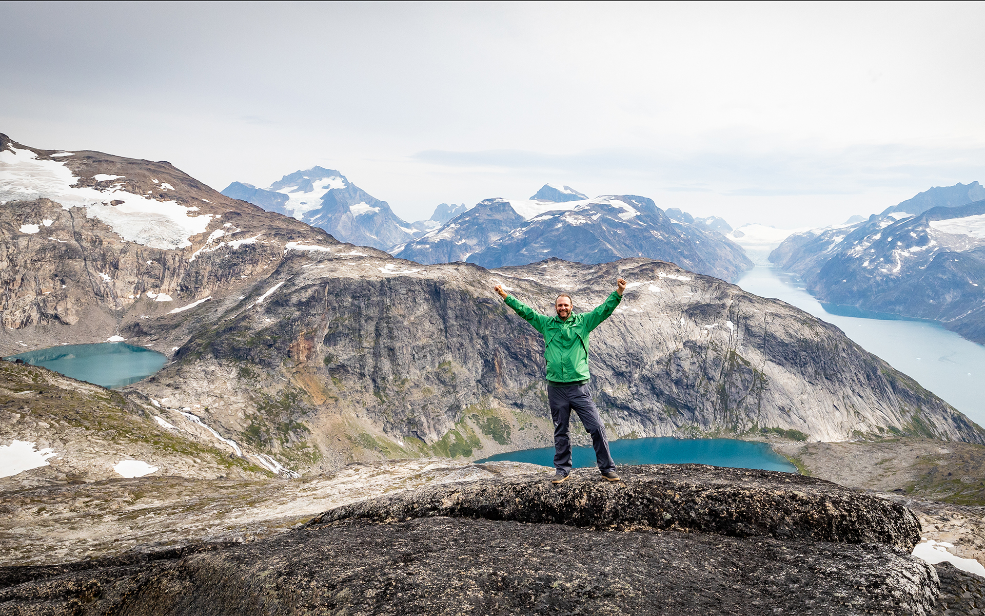 Male traveler in green parka celebrates with arms out standing atop giant mountain range with blue lakes and fjords below