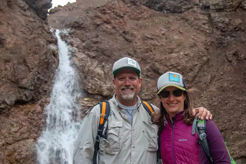 A husband and wife traveler posing in front of a rocky waterfall in Denali National Park. 