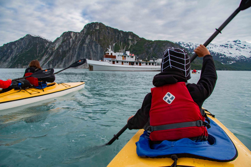 The back of a child with a red life vest and black hat kayaking toward a historic yacht over calm green water.  