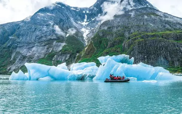 A guide on a zodiac filled with passengers in red life jackets points as he cruises the small boat among bright blue glaciers in front of a steep rocky shoreline in Alaska.