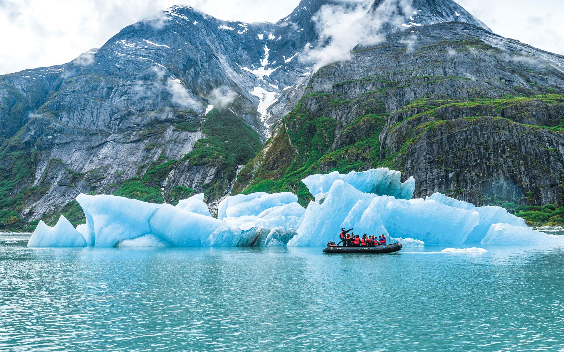 A guide on a zodiac filled with passengers in red life jackets points as he cruises the small boat among bright blue glaciers in front of a steep rocky shoreline in Alaska.