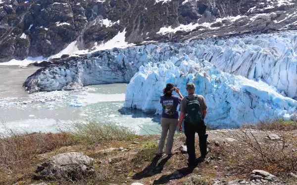 Two travelers in hats seen from behind on a hill in front of a massive blue glacier, one taking a picture with her phone and the other has a backpack on