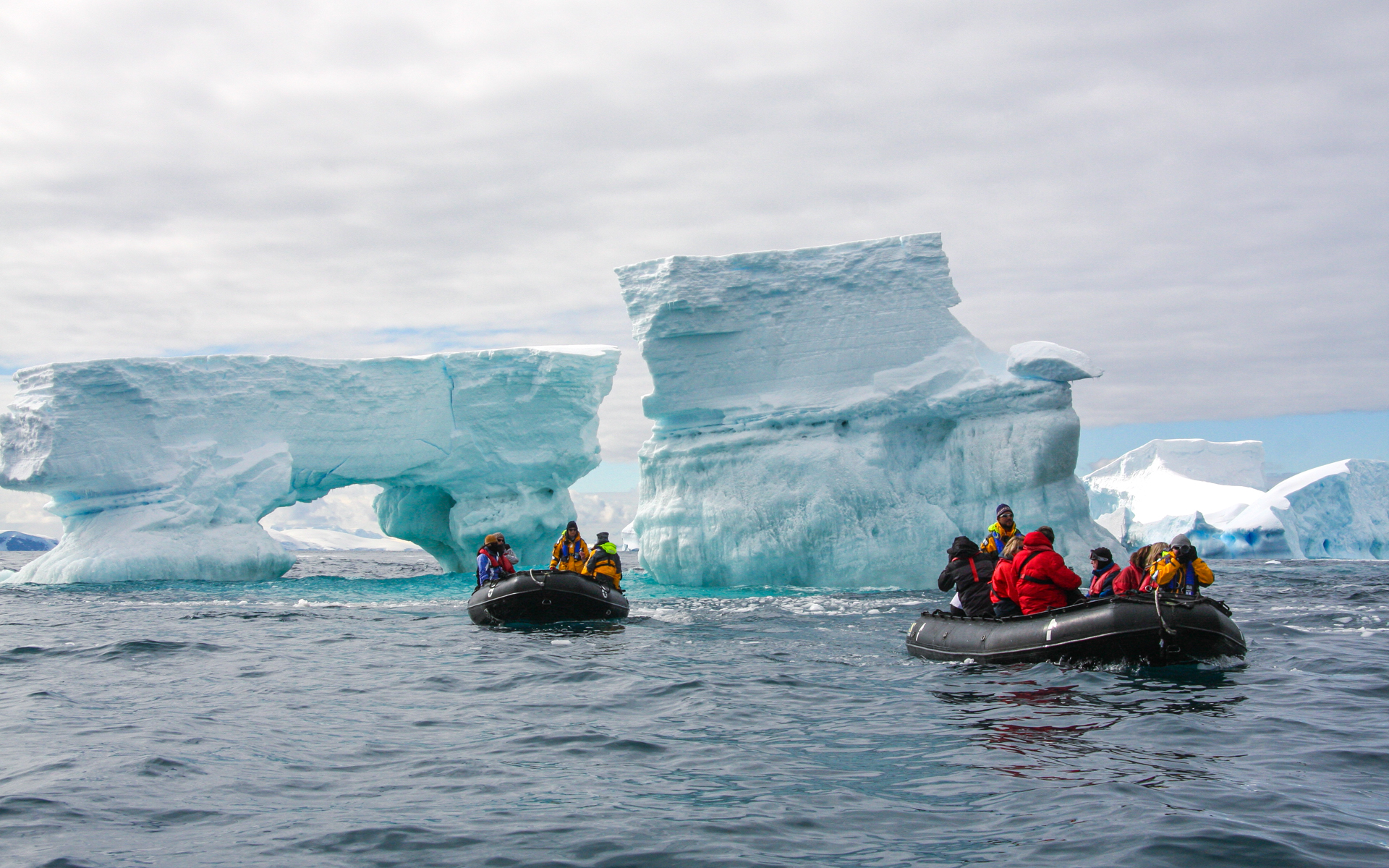 Two inflatable skiffs of travelers navigate the water while surrounded by giant teal blue and white icebergs in Antarctica