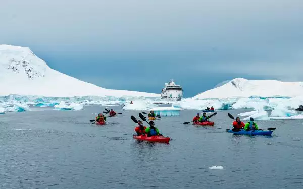 Antarctica travelers paddle brightly colored double kayaks surrounded by snowy mountains, icebergs and Greg Mortimer ship