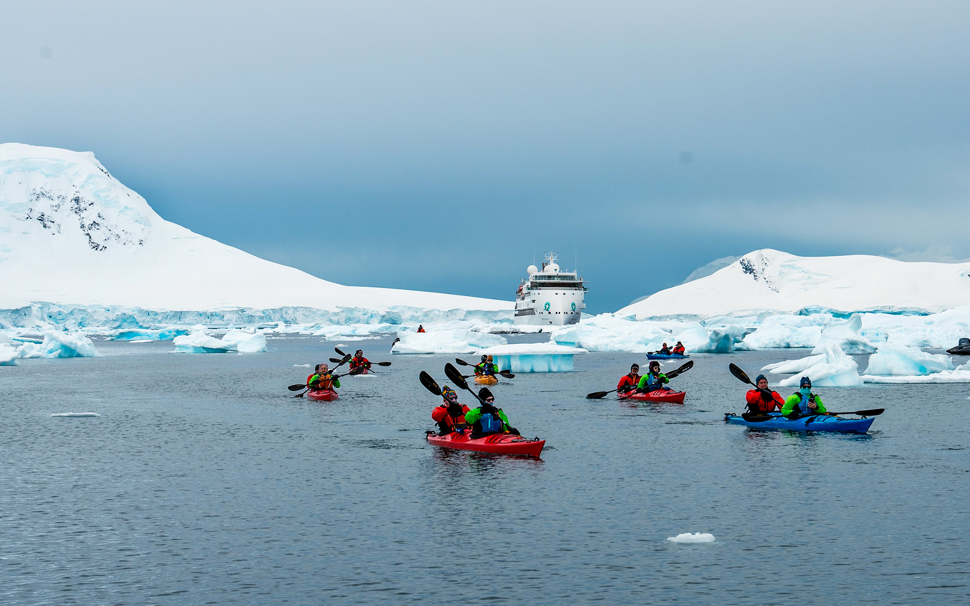 Antarctica travelers paddle brightly colored double kayaks surrounded by snowy mountains, icebergs and Greg Mortimer ship