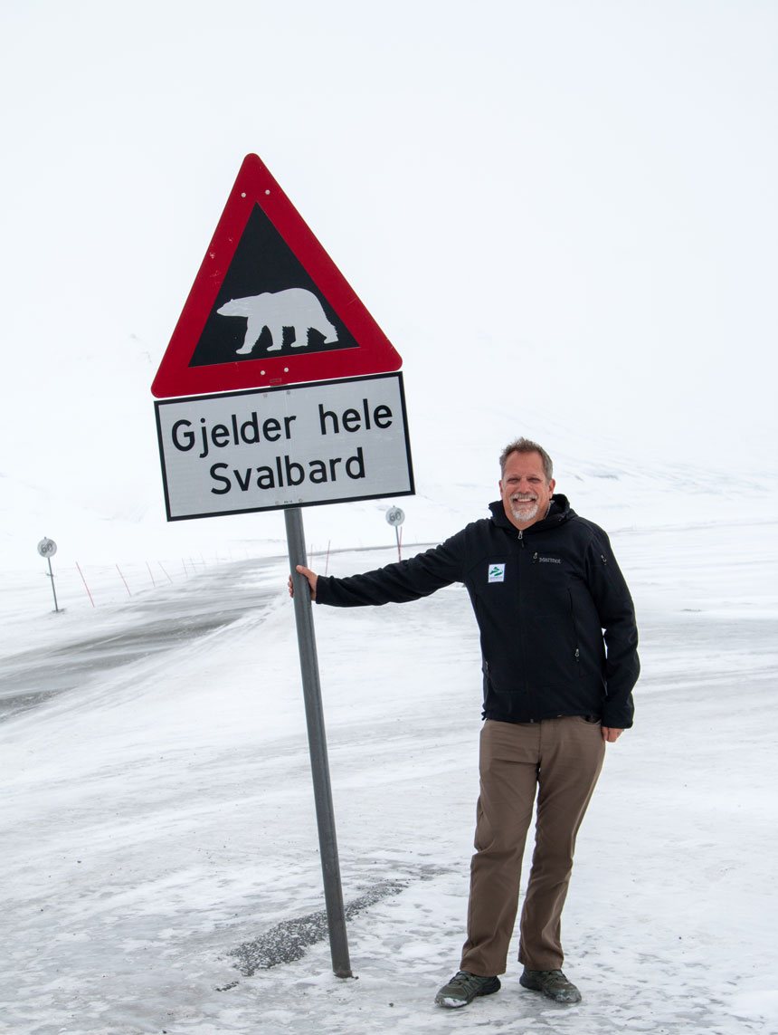 A man in a black Adventuresmith jacket with standing on ice next to a traffic sign with a polar bear on it. 