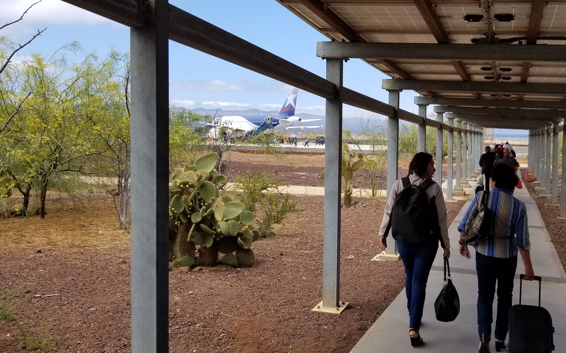 2 women walk a covered concrete path towards a loading jet plane, rolling suitcases past cacti on a sunny day in Galapagos.