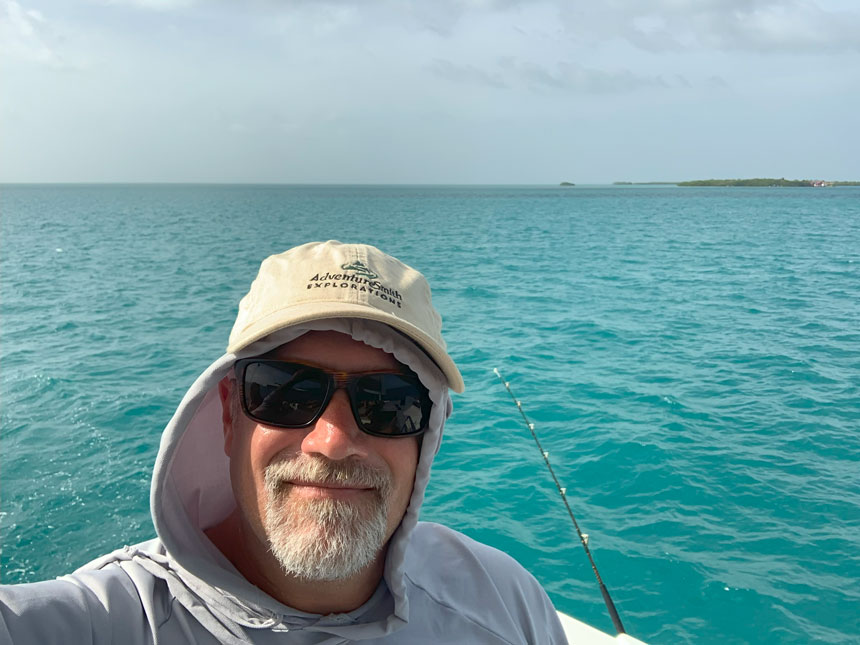 A male traveler with an AdventureSmith hat and sunglasses poses in front of a calm green ocean in Belize with a fishing pole in the water behind him.  