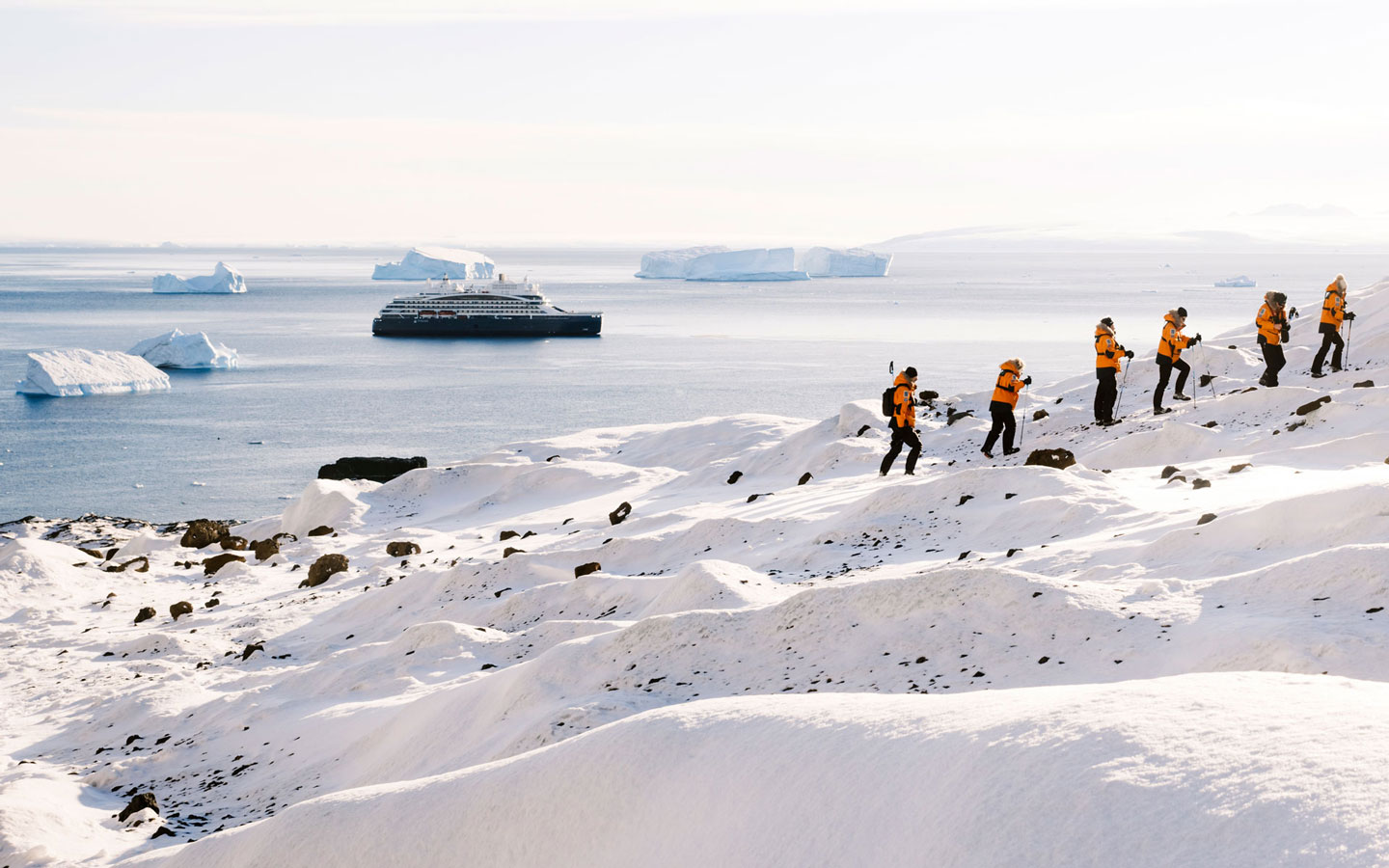 Line of polar travelers in orange coats hikes up snowy hillside in Antarctica as small ship with blue hull sits in distance.