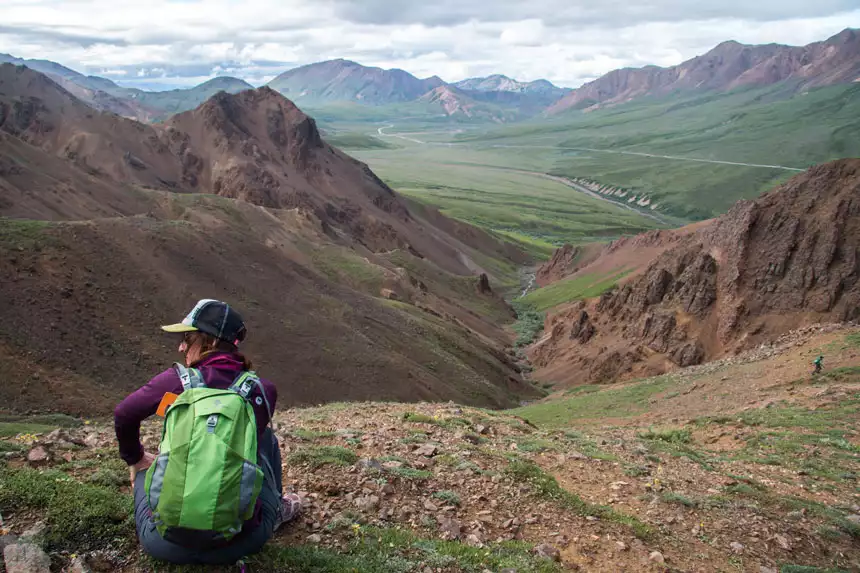 A female traveler wearing a green backpack sitting on rocks overlooking a green valley and brown mountains in Denali National Park. 