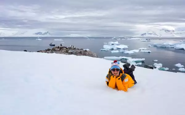 Happy female in yellow parka lays in snow with penguins, icebergs, a small ship and white Antarctica landscape behind her.