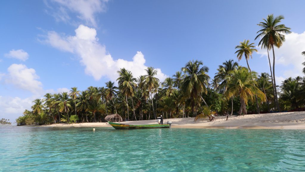 Fisherman with small colorful wood boat in turquoise water along white-sand beach with palm trees on a sunny Caribbean day.
