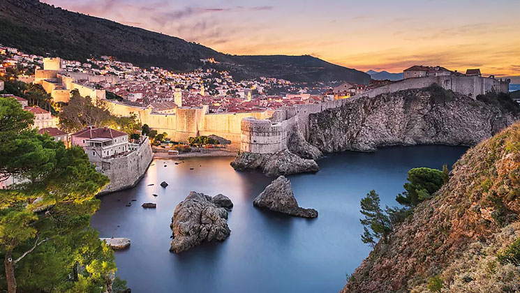 Sunset over Dubrovnik, Croatia with hillside of old stone buildings with red roofs & small inlet beach by seaside city walls.