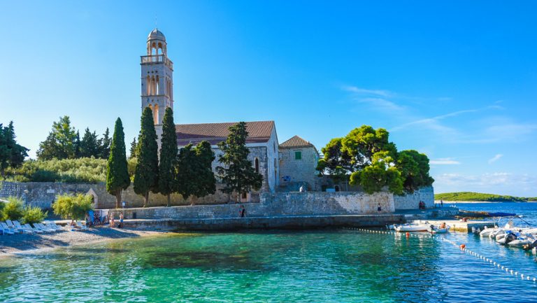 Turquoise waters by small beach & old stone building with clock tower & sea wall, seen on the Croatia Under Sail cruise.