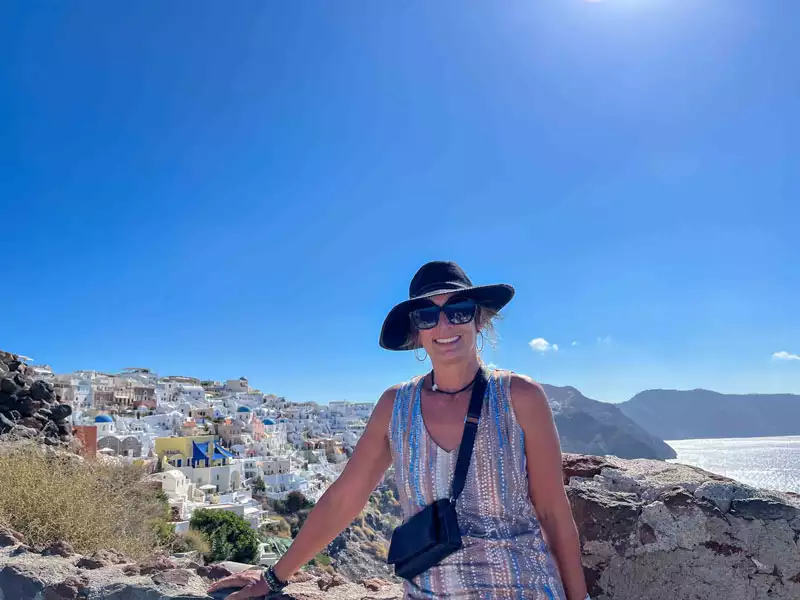 A female traveler in a hat and sunglasses overlooking a Greek village with many white houses on a cliff and the Mediterranean Sea in the background.