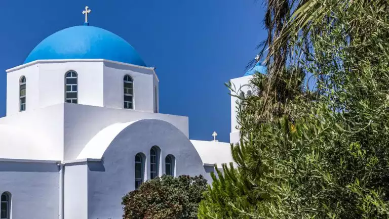 Whitewashed Greek church with blue dome & silver cross on top sits by green foliage in the sun on a Le Ponant Cyclades cruise.