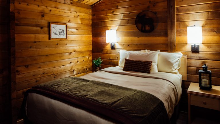 Interior of guest cabin at Denali Cabins with double bed in white bedding with green accents, bedside lights & wood walls.