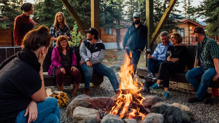 Denali Cabins adult guests sit & stand around a blazing campfire in a stone ring, drinking wine, talking & smiling.