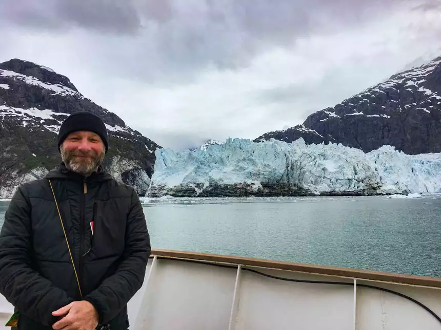 A male traveler with a black jacket poses on the deck of a small ship in front of an icy tidewater glacier. 