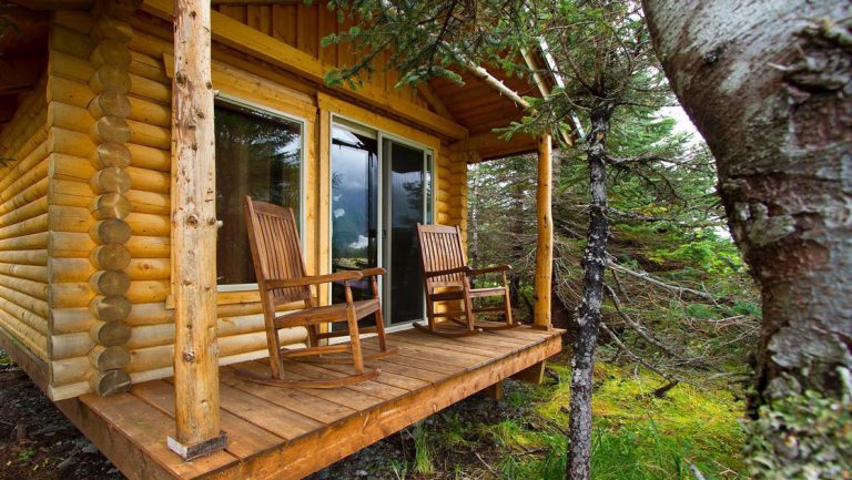 Log cabin covered porch with wooden rocking chairs, big window & sliding glass door at Kenai Fjords Glacier Lodge in Alaska.