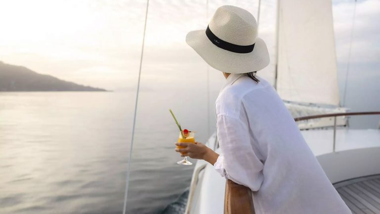 Woman in long white billowy shirt & Panama hat leans against railing & holds tropical cocktail on Sun Deck of Le Ponant ship.