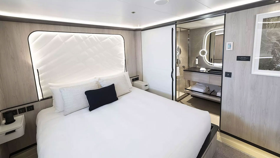 King bed in white with accent pillows & white padded headboard in beige room with pocket door to bathroom on Le Ponant ship.