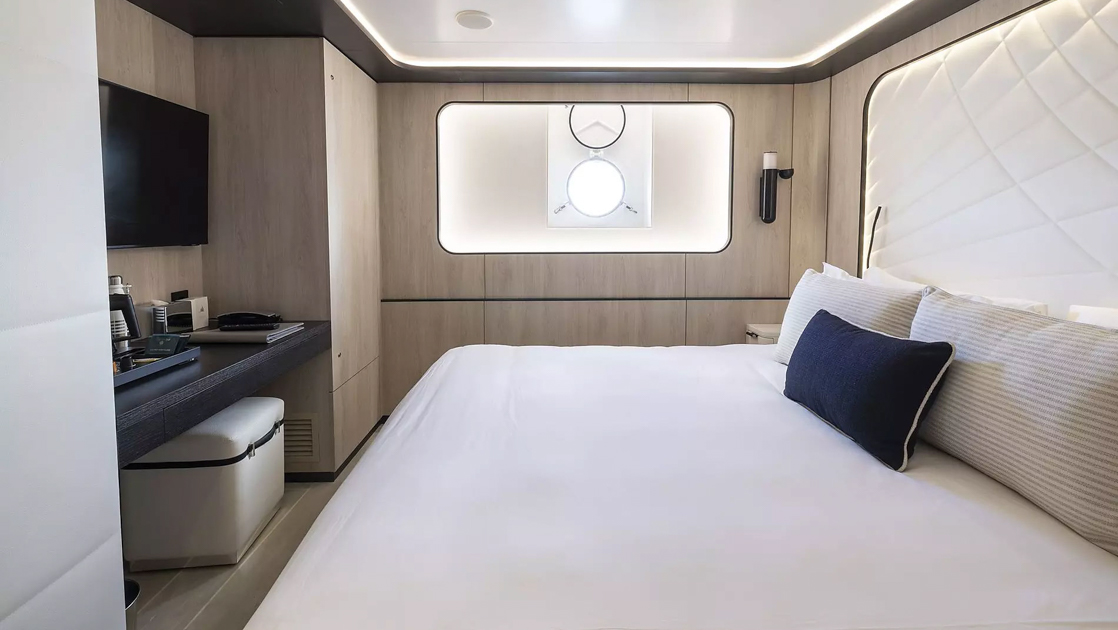 King bed in white with accent pillows & white padded headboard in beige room with porthole on Le Ponant cruise ship.