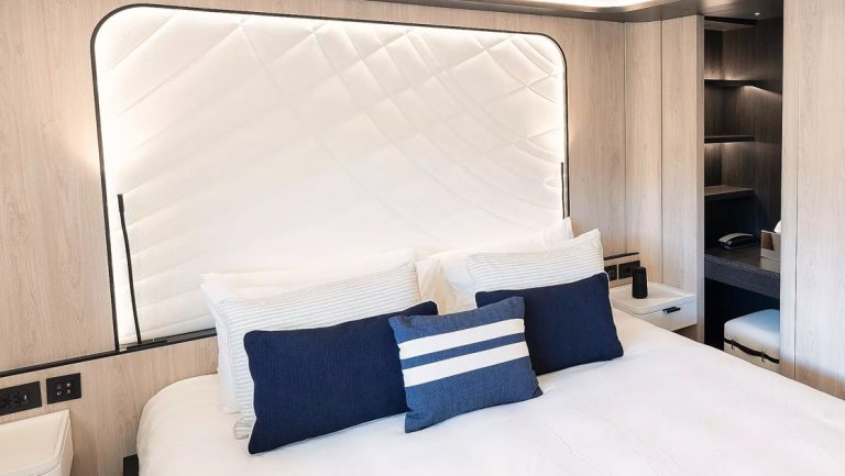King bed in white with blue accent pillows & white padded headboard in beige room on Le Ponant cruise ship.