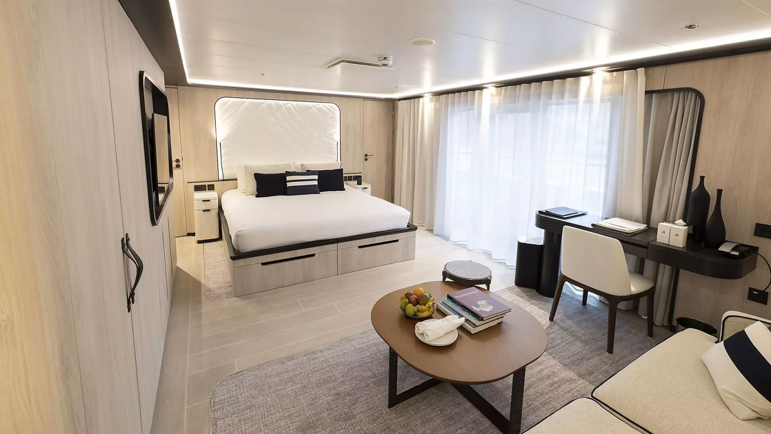King bed in white with accent pillows & white padded headboard in beige room with glass windows to balcony on Le Ponant ship.