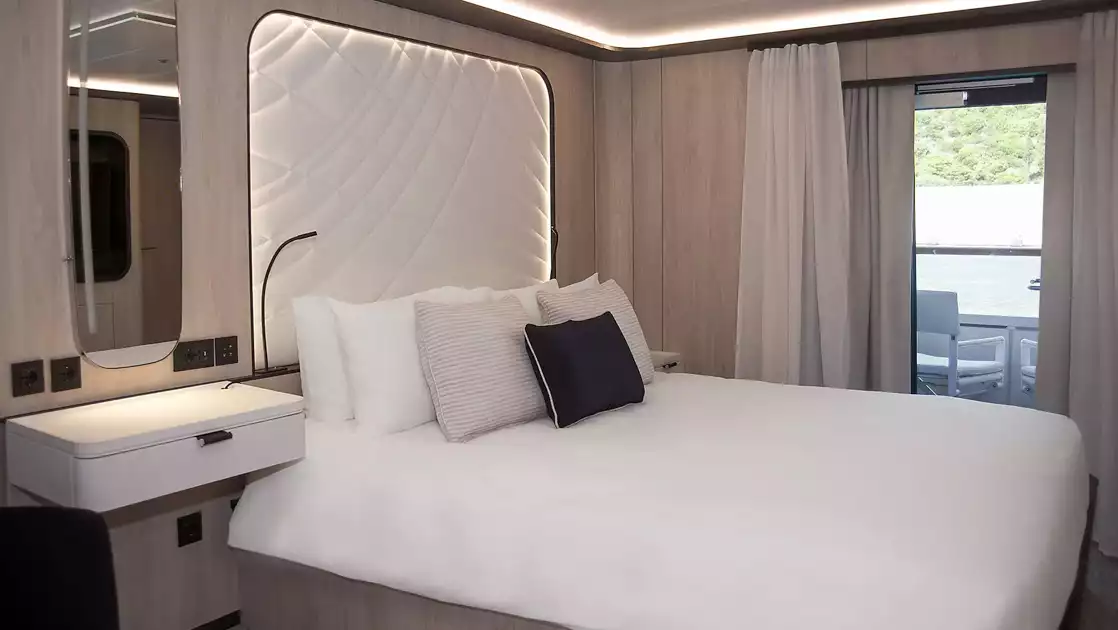 King bed in white with accent pillows & white padded headboard in beige room with glass door to balcony on Le Ponant ship.