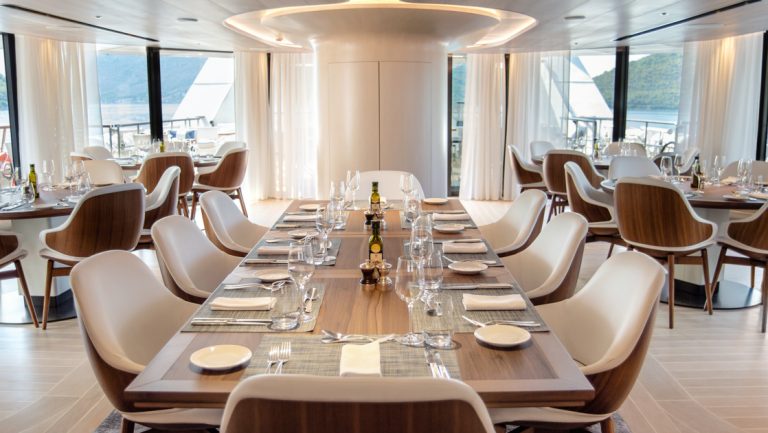 Long rectangular table & smaller round ones set for dinner on Le Ponant cruise ship with earth tone decor & padded chairs.