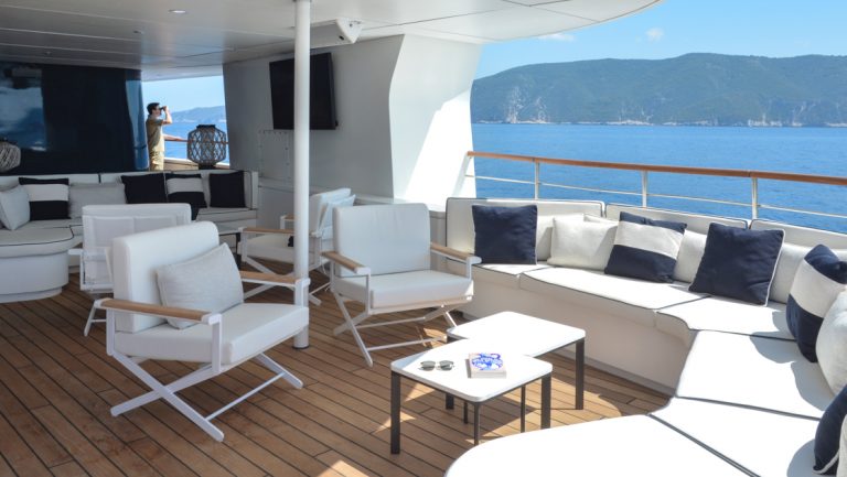 White wraparound couch & padded white chairs with small coffee tables on open-air teak deck of Le Ponant cruise ship in the sun.