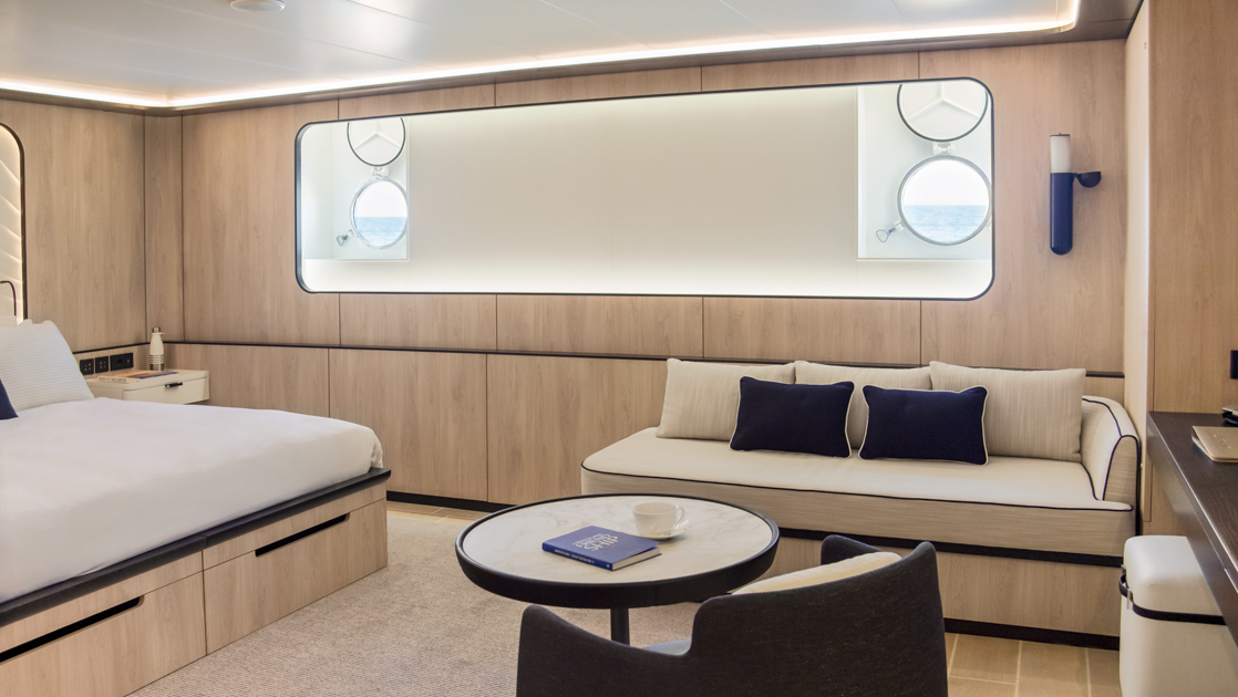 King bed in white with accent pillows & white padded headboard in beige room with 4 portholes on Le Ponant cruise ship.