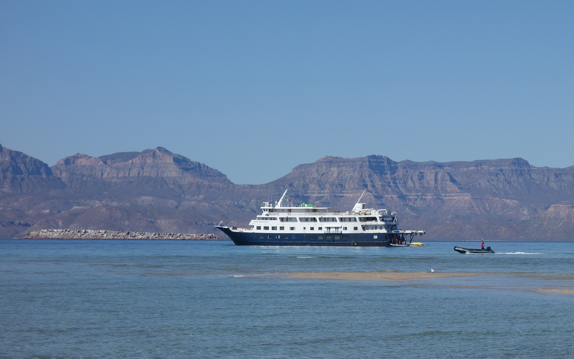 A white and blue small ship cruise seen with a zodiac behind it cruising toward the ship, with tall tan shoreline mountains behind in the distance