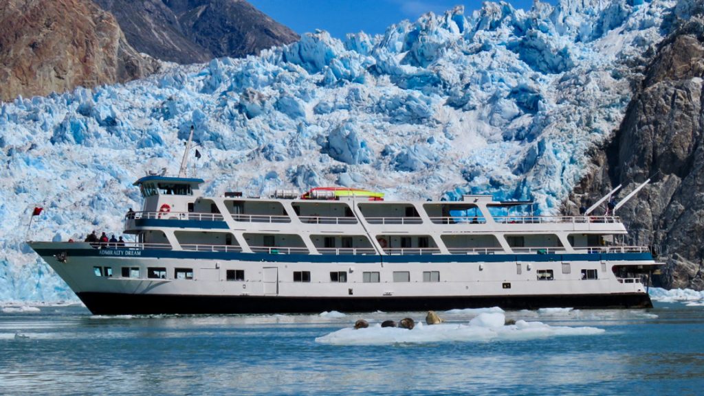 Admiralty Dream cruise ship with white decks sits parked in front of a large blue glacier on a sunny day in Alaska.