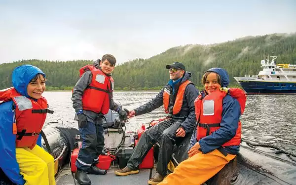 Three boy travelers in Alaska sit in a Zodiac with a guide showing them the motor, with a small ship and misty mountains behind them