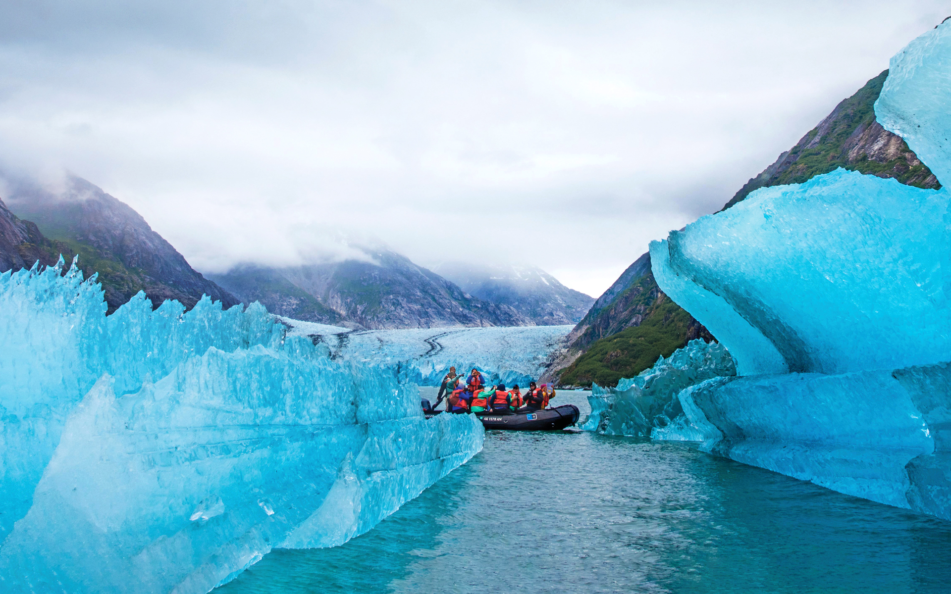 Travelers in a skiff seen dwarfed by giant blue icebergs in front of them and a larger glacier in Alaska's Inside Passage behind them.