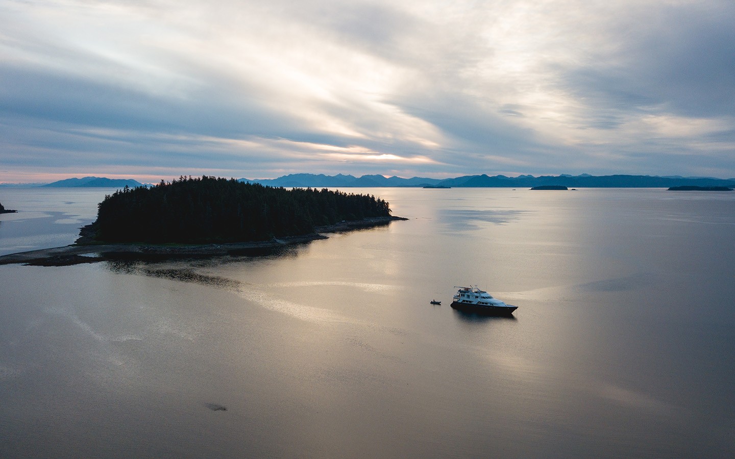A small Alaska yacht charter ship seen from above in the still waters around Admiralty Island with a grey cloudy sky at dusk