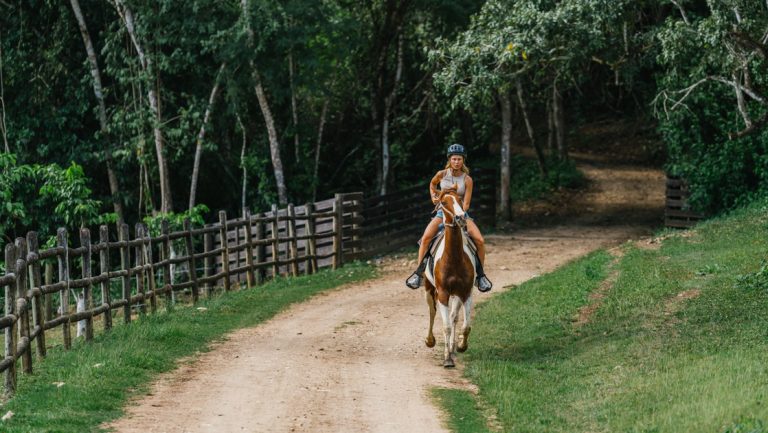 Woman rides brown & white horse on dirt road beside black wooden fence & dense forest on a Belize jungle tour.