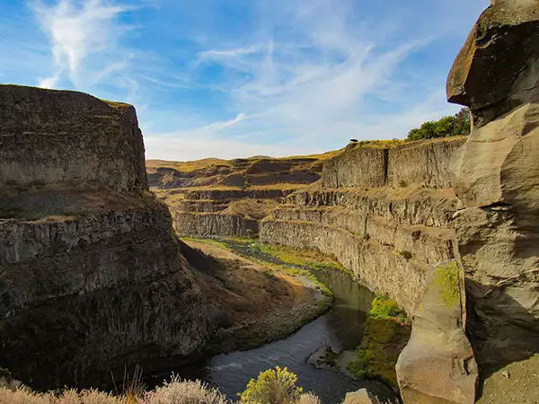Tall Columbia River canyon with crumbling tan walls carved in steps & grassy areas above & below.